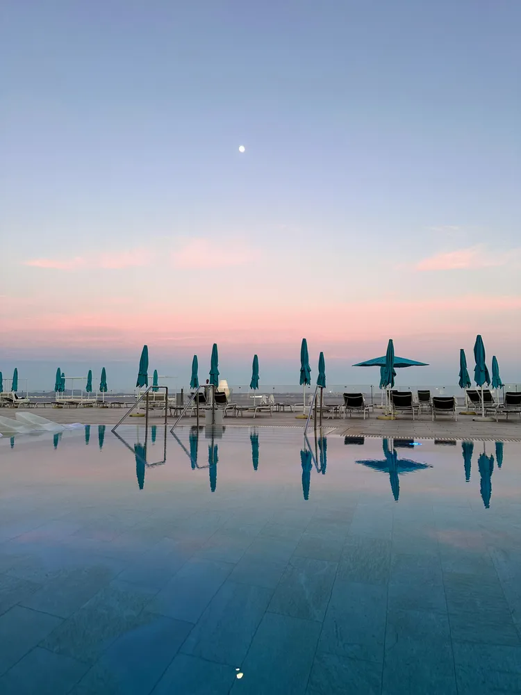 Furled parasols along a poolside at dusk, the pink sky and a low moon mirrored in still water