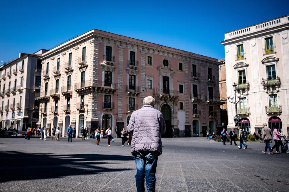 A man in a puffer jacket crossing a wide Sicilian piazza toward a pink baroque palazzo