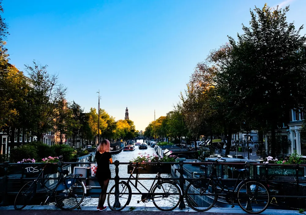A woman leaning on a canal bridge between parked bicycles, a church tower catching the last sun