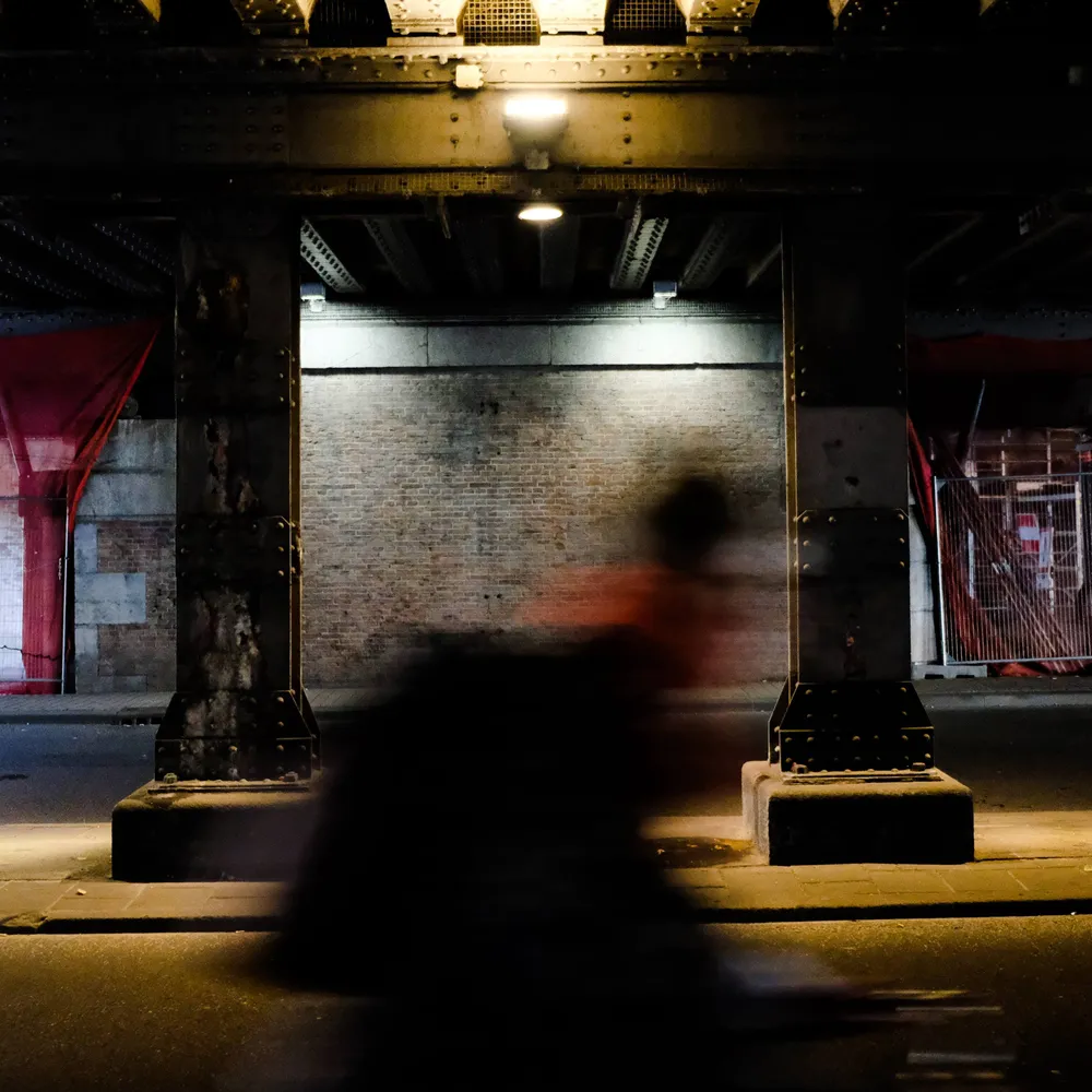A cyclist blurred to a smear of red passing a lit brick wall beneath a railway bridge at night