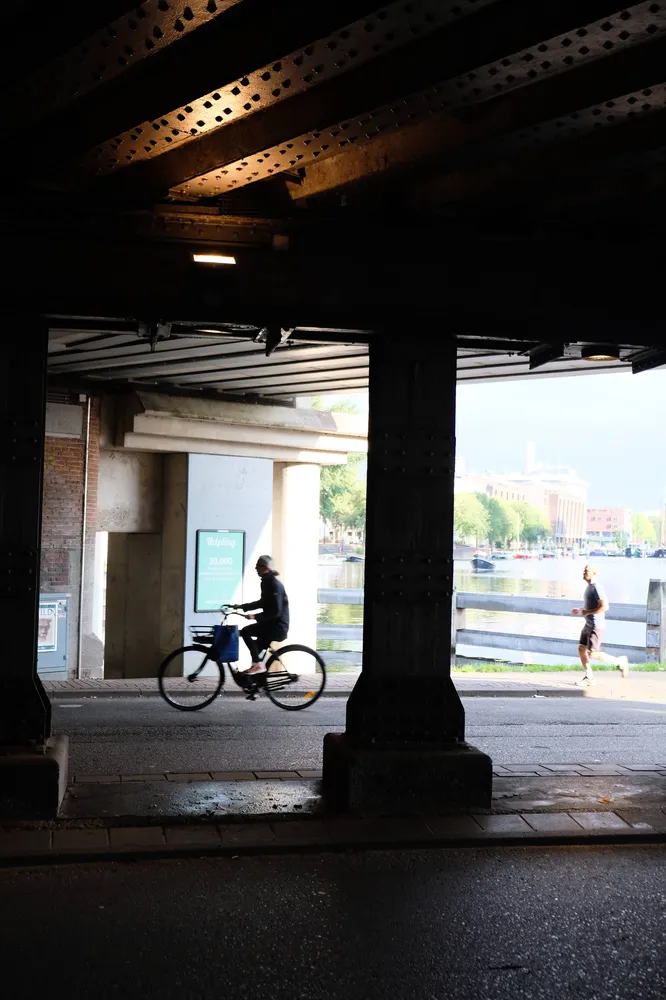 A cyclist and a runner framed by the dark ironwork of a bridge, water and trees beyond